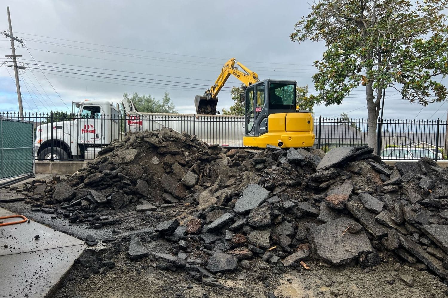 An excavator loading broken asphalt beside a D’s truck